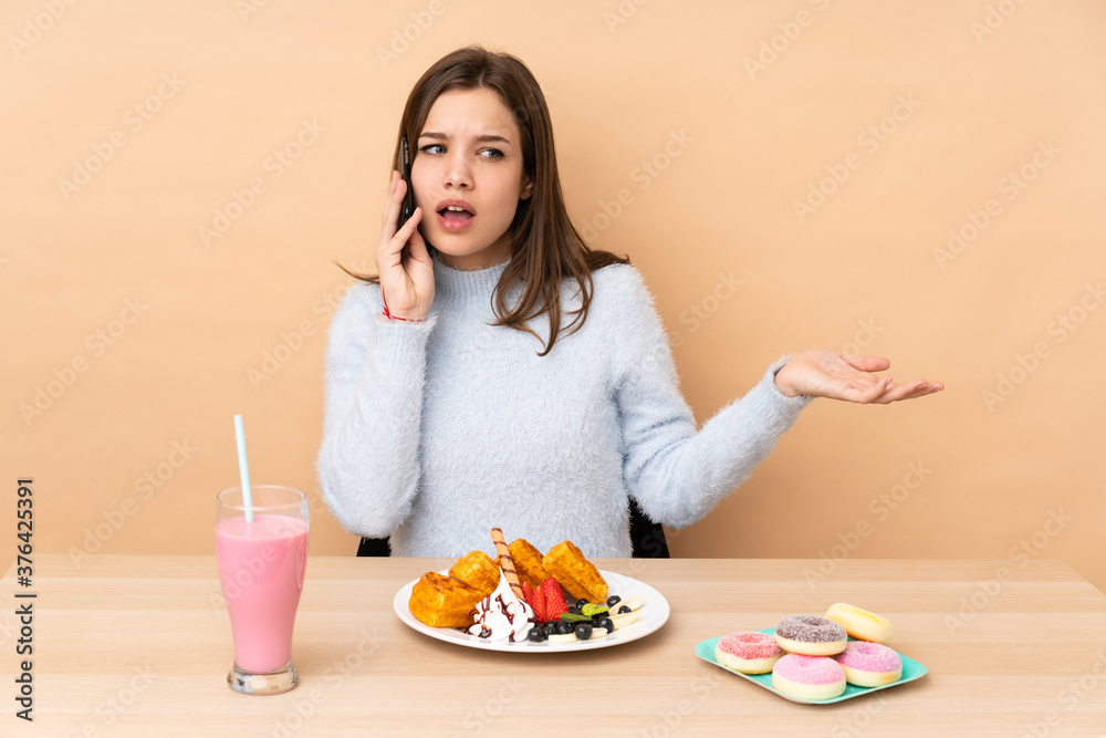 Teenager girl eating waffles isolated on beige background keeping a conversation with the mobile phone with someone Teenager girl eating waffles isolated on beige background keeping a conversation with the mobile phone with someone