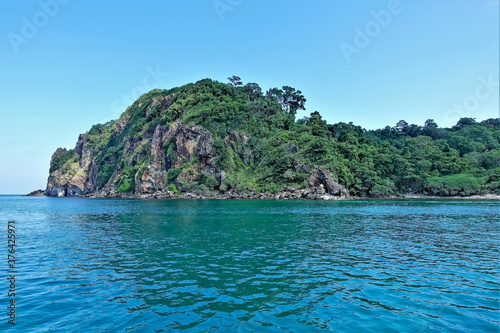 An island with green tropical vegetation rises above the aquamarine water, against the background of a blue sky. Summer in Thailand.