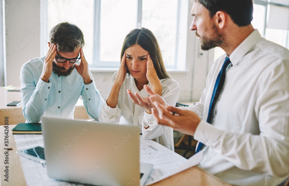 Fototapeta premium Tired office workers feeling headache during deadline reading email with bad news from colleague about project, unhappy male and female partners near desktop with blueprint and modern laptop computer
