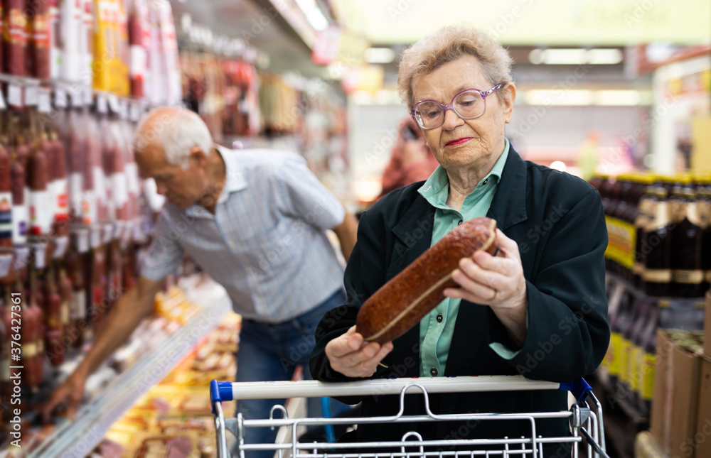 mature woman with glasses picks out salami in the meat section of the supermarket