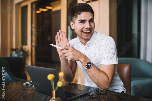 Positive male student with laptop and clipboard sitting at table in cafe raising hand
