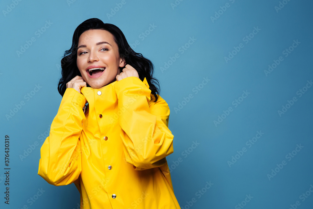 Portrait of happy trendy brunette woman smiling in yellow raincoat looking away at blank space for your advertising, sale, low prices, isolated on studio blue background. Autumn season. Portrait of happy trendy brunette woman smiling in yellow raincoat looking away at blank space for your advertising, sale, low prices, isolated on studio blue background. Autumn season.