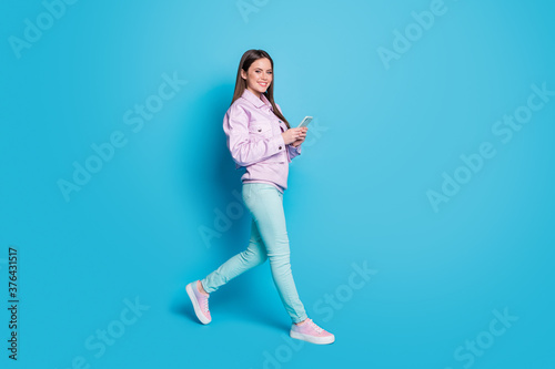 Full size profile photo of attractive lady blogger hold telephone walk street browsing phone addicted user wear casual violet jacket sweater trousers shoes isolated blue color background