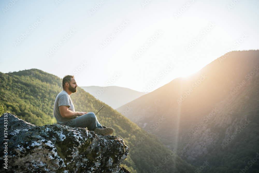 Fototapeta premium Young man sitting on a rock overlooking the mountain working with a laptop. Teleworking concept