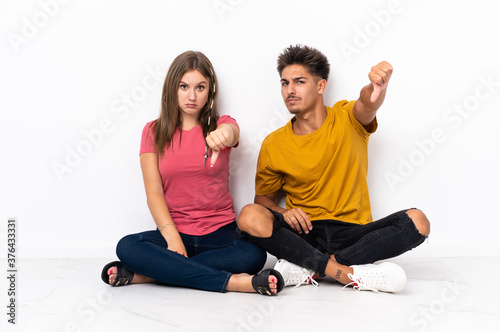 Young couple sitting on the floor isolated on white background showing thumb down with negative expression