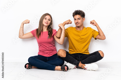 Young couple sitting on the floor isolated on white background doing strong gesture