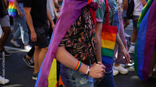 LGBT equality march. Two gays holding hands walking pride parade. Young people wearing rainbow clothes and symbols are fighting for LGBTQ+ rights. Rainbow flags, banners.