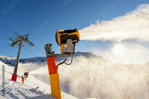 Operating artificial snow cannon near piste making snowy powder.Ski lift ropeway on hilghland alpine mountain winter resort on bright sunny day. downhill slopes with people enjoy sport activities photo