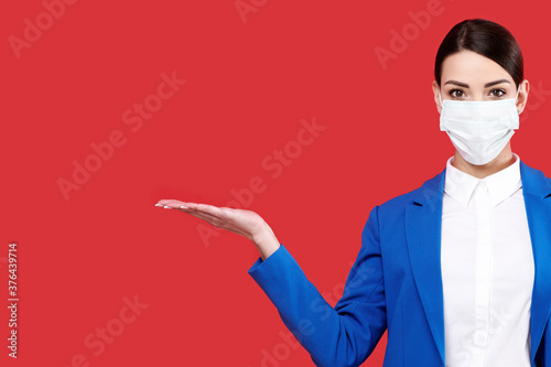 Young caucasian business woman wearing medical protective mask and blue suit holding something on red isolated background