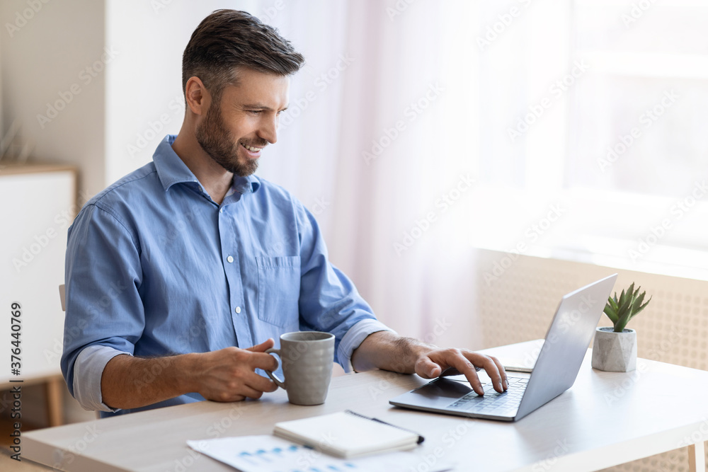 Home Office. Smiling Freelancer Man Checking Emails On Laptop And Drinking Coffee Home Office. Smiling Freelancer Man Checking Emails On Laptop And Drinking Coffee