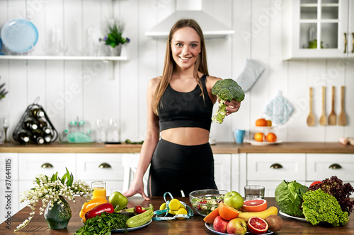 A sporty girl holds broccoli and stands in the kitchen near a table with fruits and other useful foods. Healthy eating concept