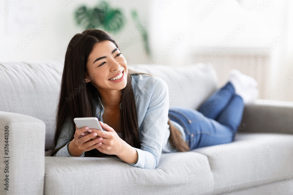 Carefree Asian Girl Enjoying Rest On Comfortable Sofa With Smartphone, Sincerely Smiling Carefree Asian Girl Enjoying Rest On Comfortable Sofa With Smartphone, Sincerely Smiling