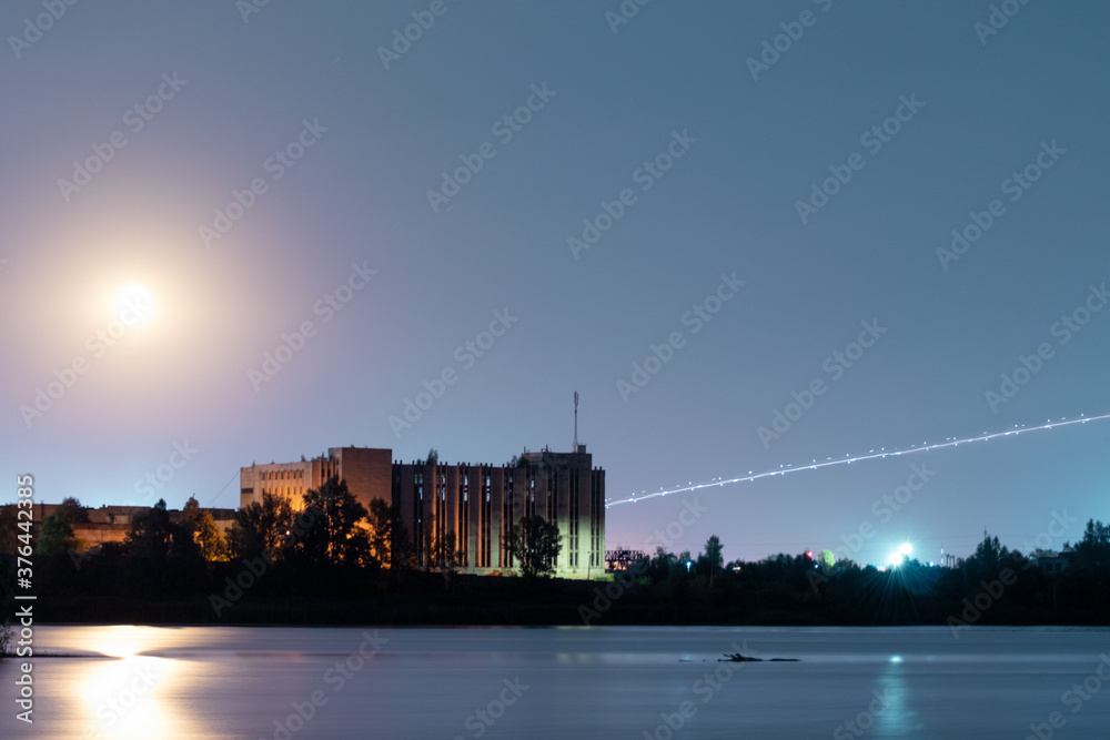 Bright moon shining on dark blue night sky above city lake water surface with reflections. Nighttime with large moon and airplane lights in long exposure