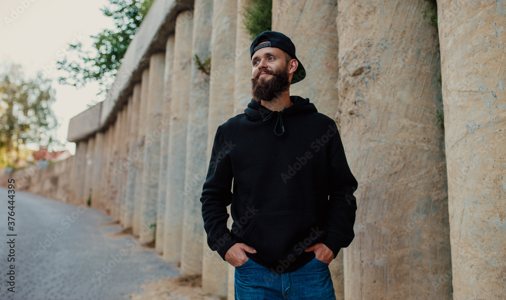 City portrait of handsome hipster guy with beard wearing black blank hoodie or sweatshirt with space for your logo or design. Mockup for print City portrait of handsome hipster guy with beard wearing black blank hoodie or sweatshirt with space for your logo or design. Mockup for print