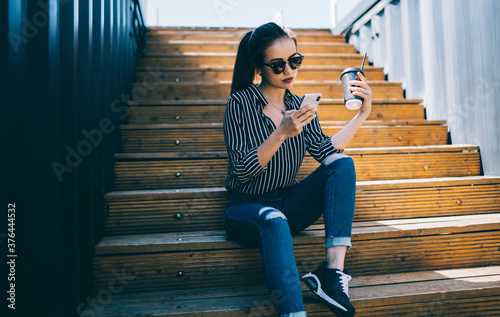 Millennial blogger in stylish sunglasses writing email message for send during mobility browsing, Caucasian hipster girl with coffee to go resting at urban setting using cellular for creating content © BullRun