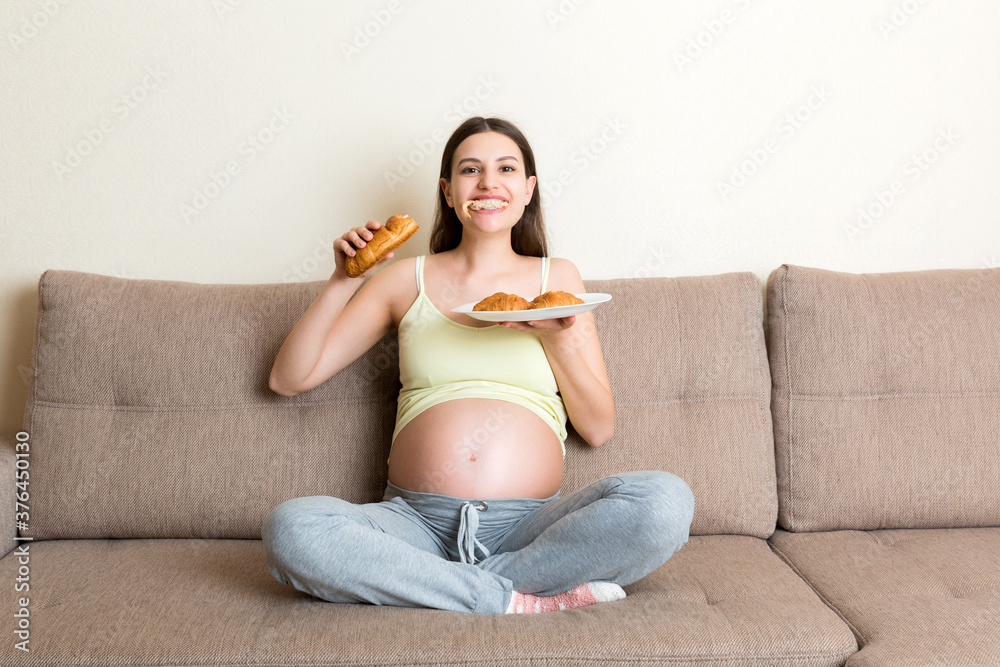 Fototapeta premium Young pregnant woman enjoys eating croissant on the sofa. Unhealthy pastry during pregnancy concept