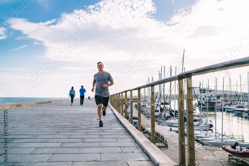 Smiling caucasian male jogger enjoying training on coastline feeling energy and vitality for keeping perfect body shape, 20s cheerful young man running on street on morning cardio workout