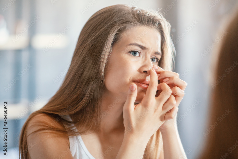 Displeased young woman squeezing acne on her nose while looking in mirror indoors Displeased young woman squeezing acne on her nose while looking in mirror indoors