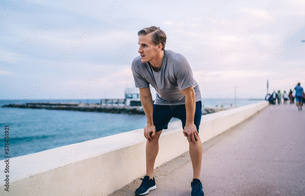 Young caucasian male jogger breathing on pause during running training legs on morning outdoor workout,thoughtful sportsman looking at water standing on quay resting on training keeping healthy body Young caucasian male jogger breathing on pause during running training legs on morning outdoor workout,thoughtful sportsman looking at water standing on quay resting on training keeping healthy body