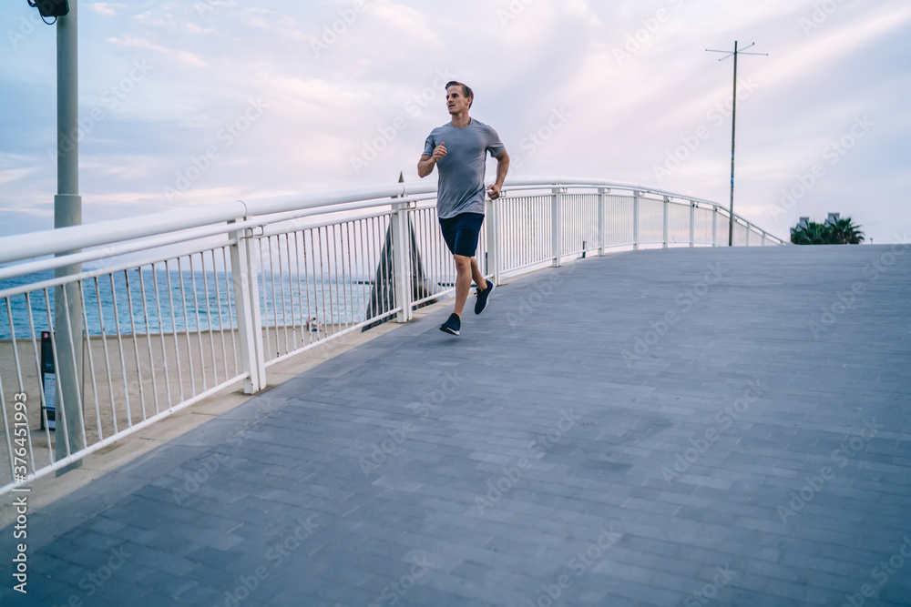 Young caucasian male athlete training outdoors keeping perfect body share and muscular strength, slim handsome man jogging on bridge on morning workout for keeping flexibility and physical power