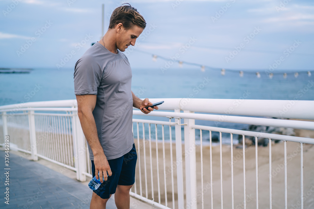 Handsome male athlete with muscular figure reading notification on mobile phone after training outdoors, 20s hipster guy keeping healthy lifestyle checking results for cardio workout on street