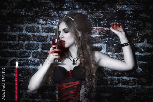 A vampire woman looks at a bloody flower. A glass of blood. Brick wall. 