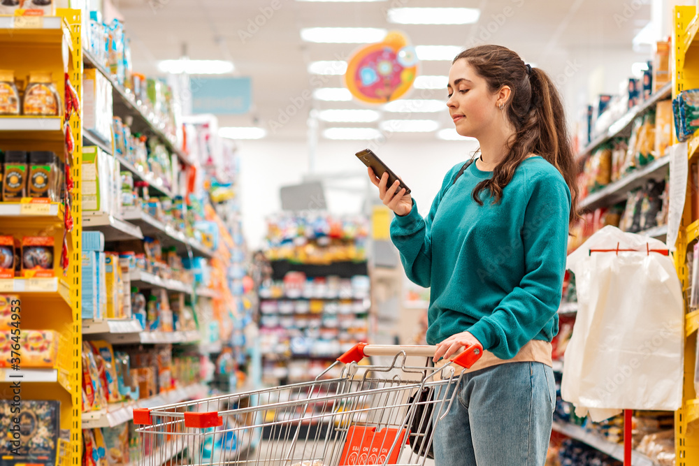 A young pretty Caucasian woman in casual clothes, pushing a grocery cart and using a mobile phone. Copy space. The concept of shopping and buying food and communication