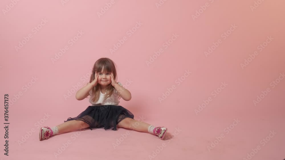 A cute little girl sits on the floor and indulges, smiles grimaces. Beautiful bright festive outfit. Pink background. Stock-Video | Adobe Stock 
