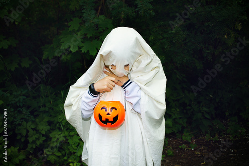 Child in halloween ghost costume looking down at basket of pumpkin in the parkt among green trees. Halloween celebration  masquerade  trick or treat.