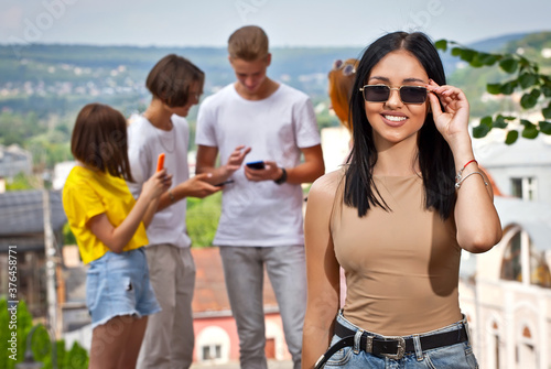 Friends drinking coffee on the background of the city. Boys and girls are smiling. Teenagers on a sunny day in the square. Young people looking at a smartphone.