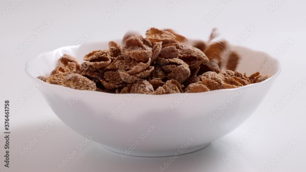 Natural whole grain wheat flakes are falling to white bowl. Healthy breakfast concept. Plate with multigrain flakes on white background Natural whole grain wheat flakes are falling to white bowl. Healthy breakfast concept. Plate with multigrain flakes on white background