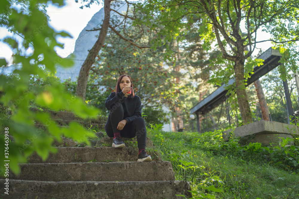 Low angle of young female traveler in activewear sitting on old stone stairs and drinking coffee while resting during walk in green botanical park