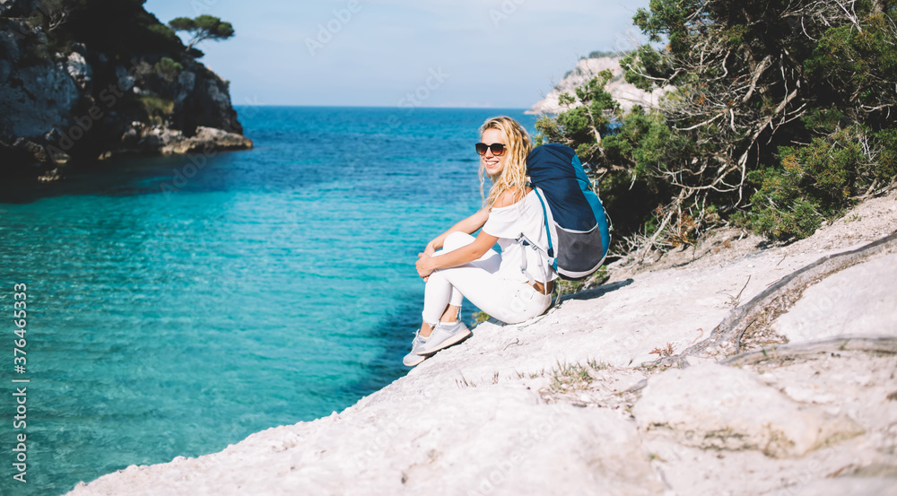 Portrait of cheerful female tourist resting at rocky abyss satisfied with getaway travel vacations for recreation during Spanish visiting, happy woman with touristic backpack smiling at camera