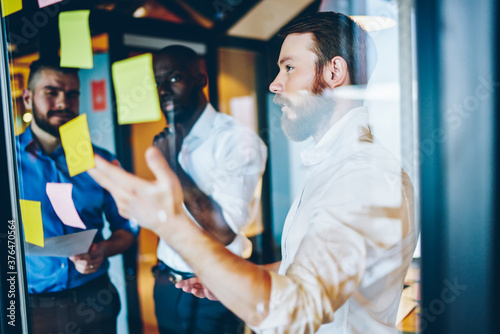 Multiracial male employees discussing business strategy during brainstorming collaboration, diverse colleagues analyzing memo notes on glued stickers talking about exchange cooperation in office