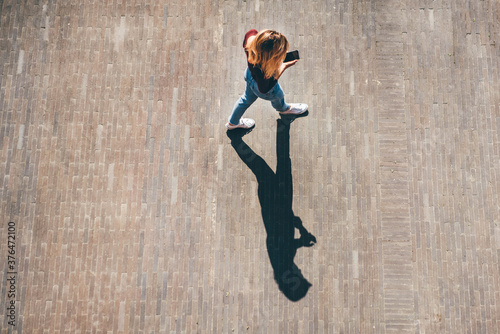 Young blonde woman in casual clothes walks across empty paved city square looking at modern smartphone screen on sunny summer day upper view.