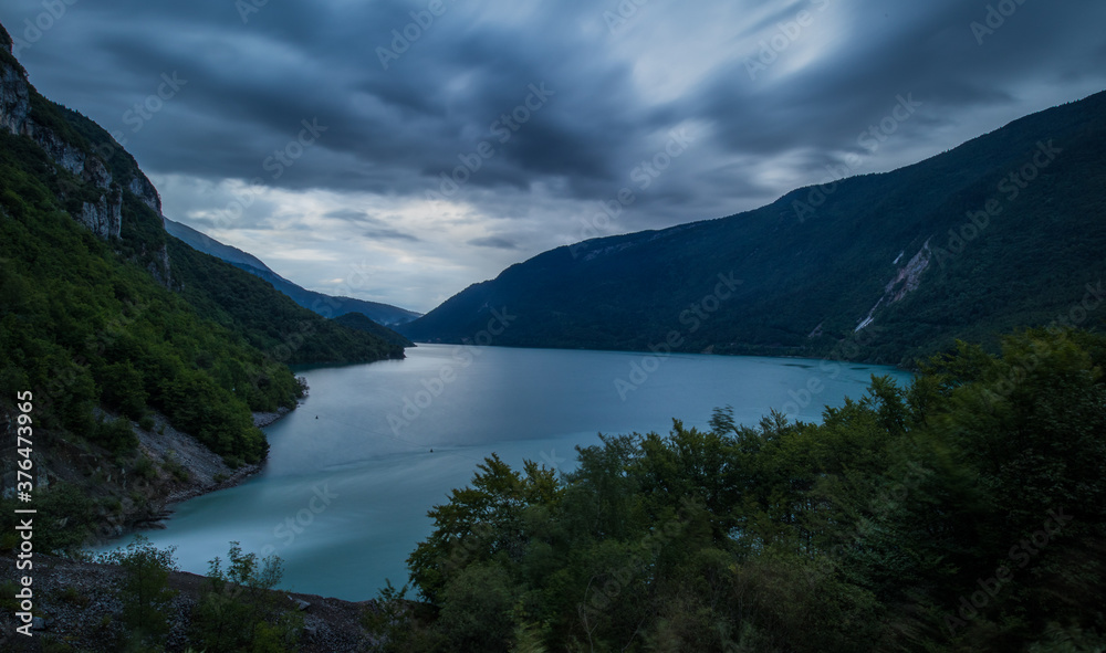 Evening panorama of Molveno lake with visible overflow waterfall of the power plant with wild white water falling into the lake. Long exposure of clouds