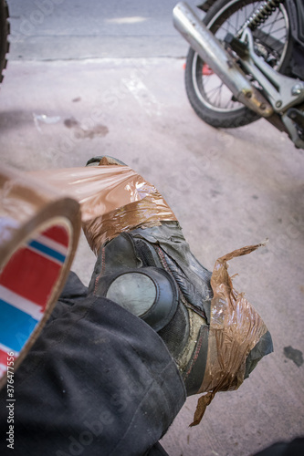 A person taping up his broken or used leather motorcycle boots with brown tape or sellotape in order to lengthen their lifespan. Motorcycle in the background. photo