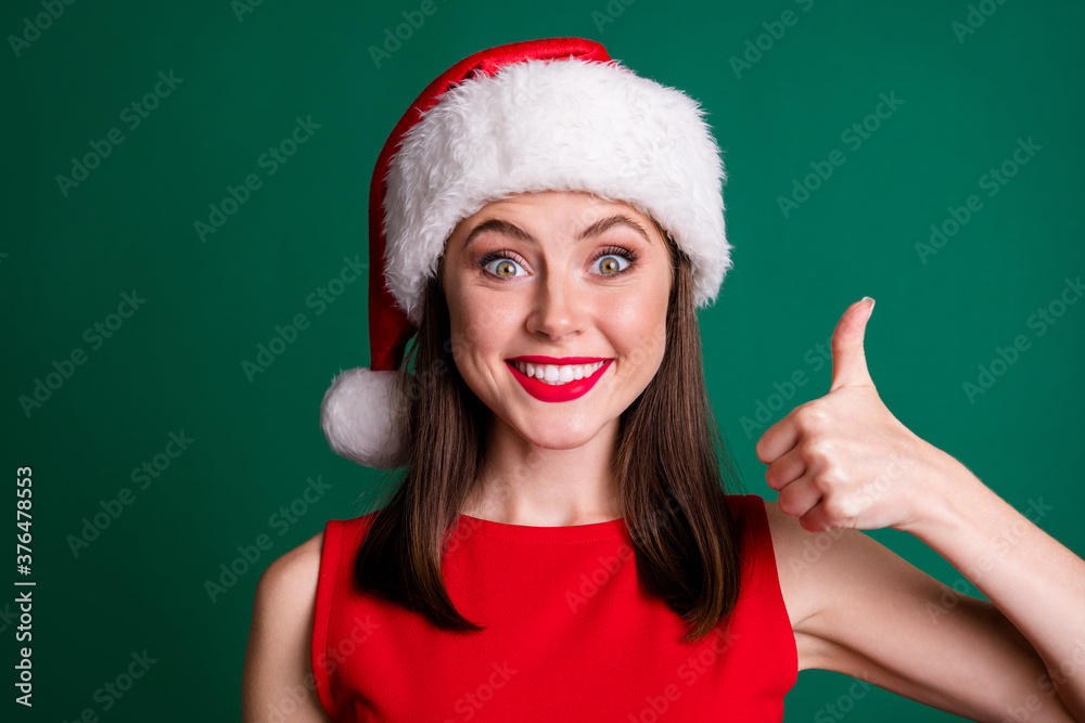 Close-up portrait of her she nice attractive charming lovely content funky ecstatic cheerful cheery girl wearing Santa cap showing thumbup cool isolated over green color background