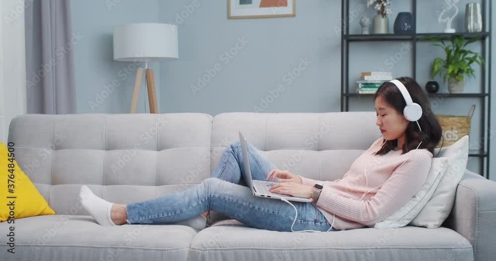 Pretty Asian girl lying on couch and using laptop computer in living room. Joyful young woman in headphones listening to music, chatting with friend. Social media, Internet, leisure time concept. Pretty Asian girl lying on couch and using laptop computer in living room. Joyful young woman in headphones listening to music, chatting with friend. Social media, Internet, leisure time concept.