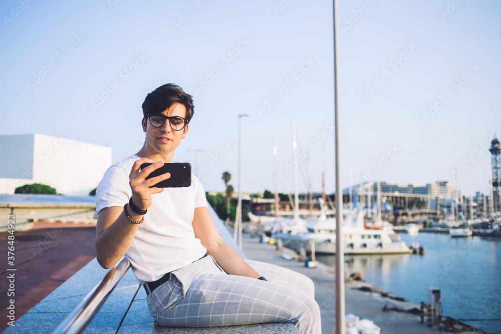 Handsome caucasian male influencer taking photo during vacation trip resting in marine port with boats, young trendy dressed male travel blogger shooting video from journey using smartphone app