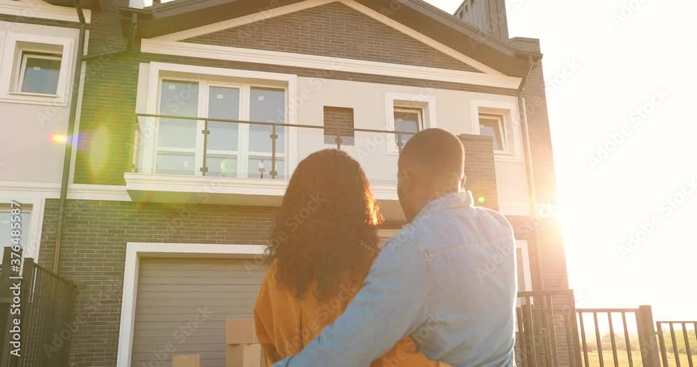 Rear on African American young just-married couple standing at yard of house, hugging and observing second floor. Back view on man and woman in hugs moving in new home. Rear on African American young just-married couple standing at yard of house, hugging and observing second floor. Back view on man and woman in hugs moving in new home.