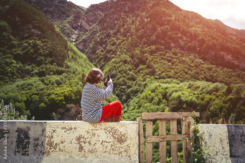 Naklejka premium A brown-haired girl in a striped black and white sweater makes a photo of the mountains on a smartphone. North Ossetia