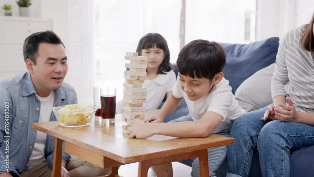 Asian family playing board game with wooden tower together at home. Son is pulls out a brick while sister, father and mother is looking and cheering.Weekend activity with entertainment at home concept Asian family playing board game with wooden tower together at home. Son is pulls out a brick while sister, father and mother is looking and cheering.Weekend activity with entertainment at home concept