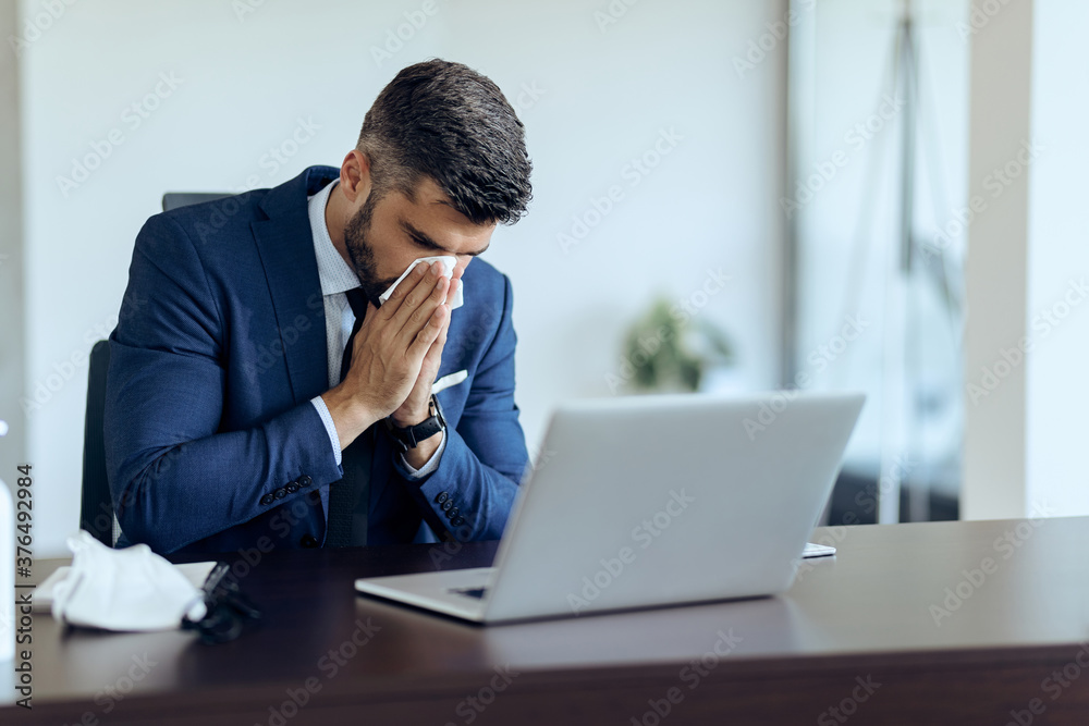 Sick businessman sneezing in a tissue at his office desk. Sick businessman sneezing in a tissue at his office desk.