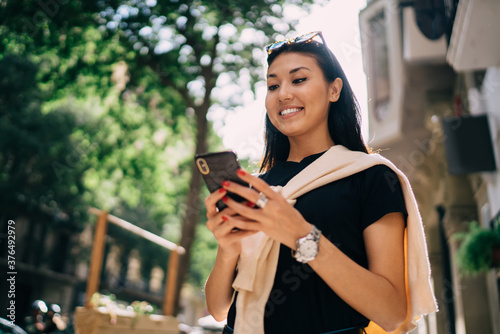 Cheerful asian milnielan woman happy about getting notification from banking app on smartphone, positive hipster female 20s using mobile phone for communication on free time outdoors on street
