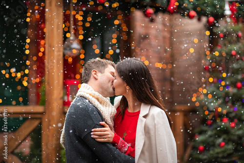 Young man and pretty woman in winter sweaters kissing under the snow in the back yard. Christmas background.
