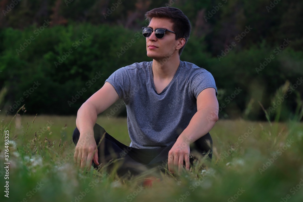 Caucasian Man with Sunglasses Sitting on Meadow and Posing. Young Adult Relaxing in the Nature in Czech Republic. Caucasian Man with Sunglasses Sitting on Meadow and Posing. Young Adult Relaxing in the Nature in Czech Republic.