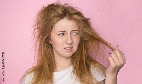 The concept of dry lifeless hair. A woman on a pink background holds her disheveled, tangled hair and looks at it with a discontented expression.
