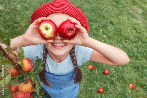 Apples for Children. Eco Organic Apple Harvest. Little girl in red hat holds two apples near the eyes on grass background.  © Maryana