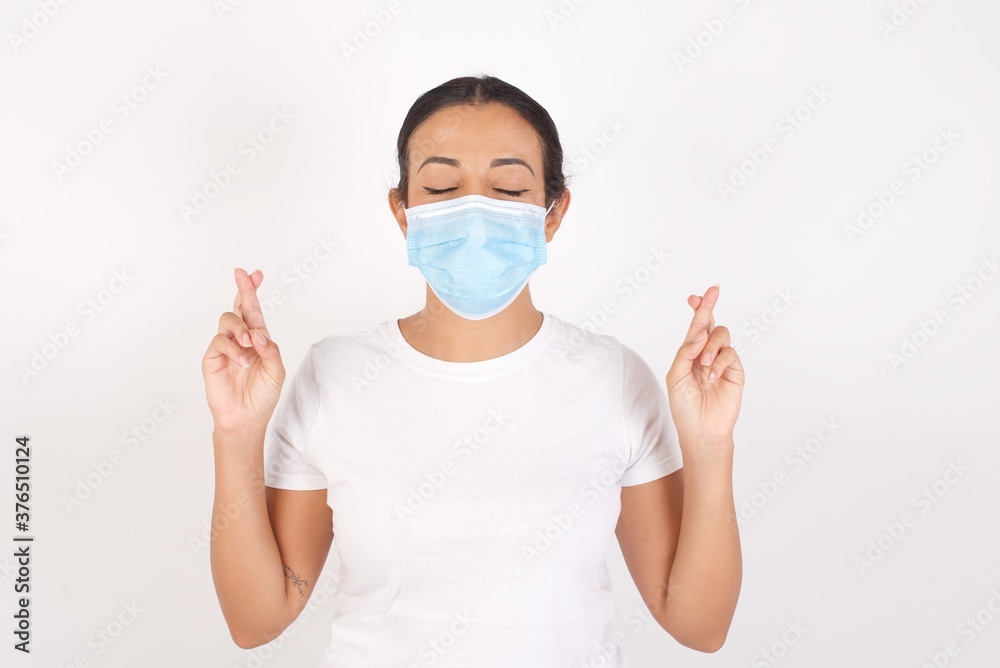 Young arab woman wearing medical mask standing over isolated white background has big hope, crosses fingers, believes in good fortune, smiles broadly. People and wish concept